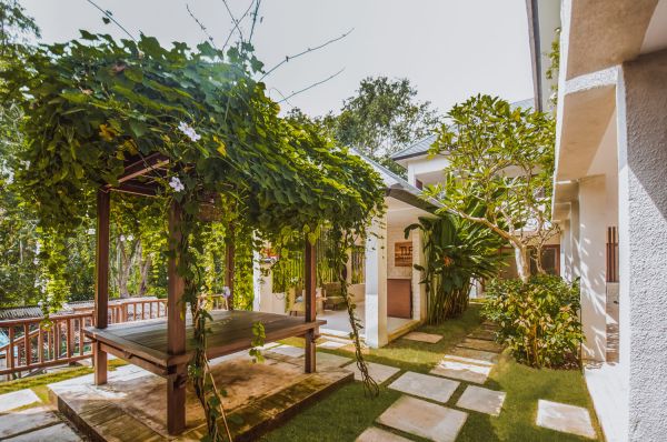 Lush tropical pergola covered in vines with wooden bench in serene garden courtyard, white Balinese-style buildings and greenery. - Bali Villas